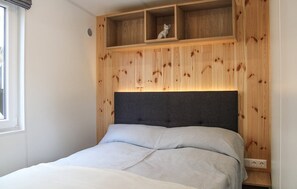A bedroom with a bed, a headboard, a shelf with a cat, and a window.