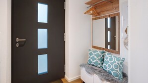 Una puerta oscura con cuatro ventanas cuadradas, un perchero de madera, un espejo y cojines estampados en un banco blanco.