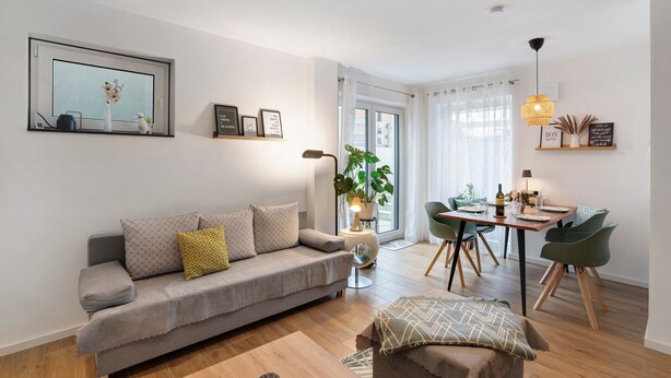 Una sala de estar moderna con un sofá gris, una mesa de comedor con sillas, una planta en maceta y una ventana con cortinas.