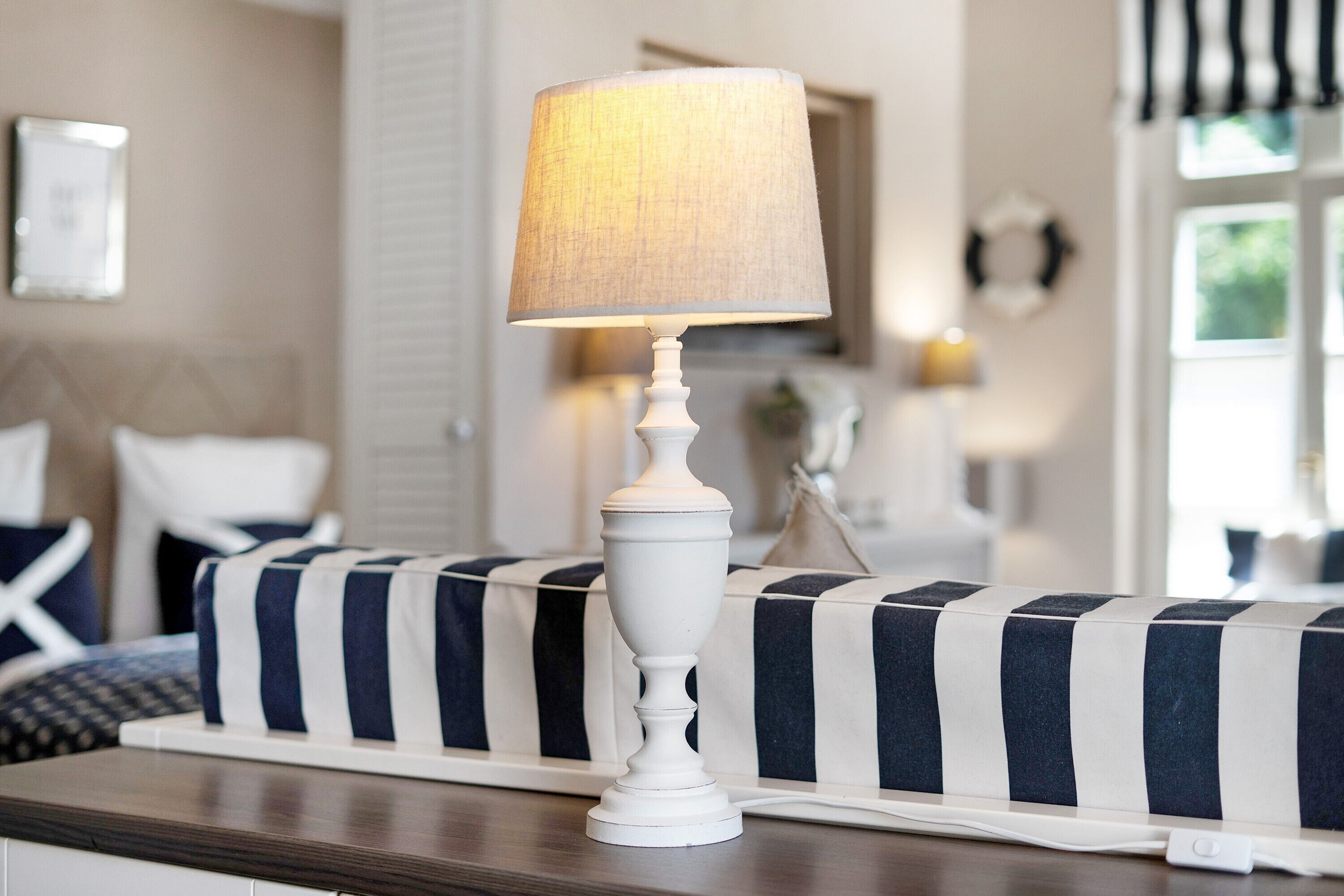 A white table lamp with a cylindrical shade on a wooden table, a striped sofa, and a bed with white bedding in a room with a neutral color scheme.