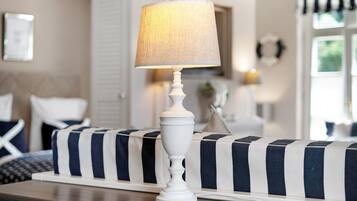 A white table lamp with a cylindrical shade on a wooden table, a striped sofa, and a bed with white bedding in a room with a neutral color scheme.
