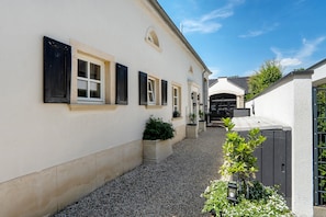 Ein schmaler Gang mit einem Gebäude, das eine Reihe von Fenstern mit Fensterläden, einen Kiesweg und Topfpflanzen zeigt.