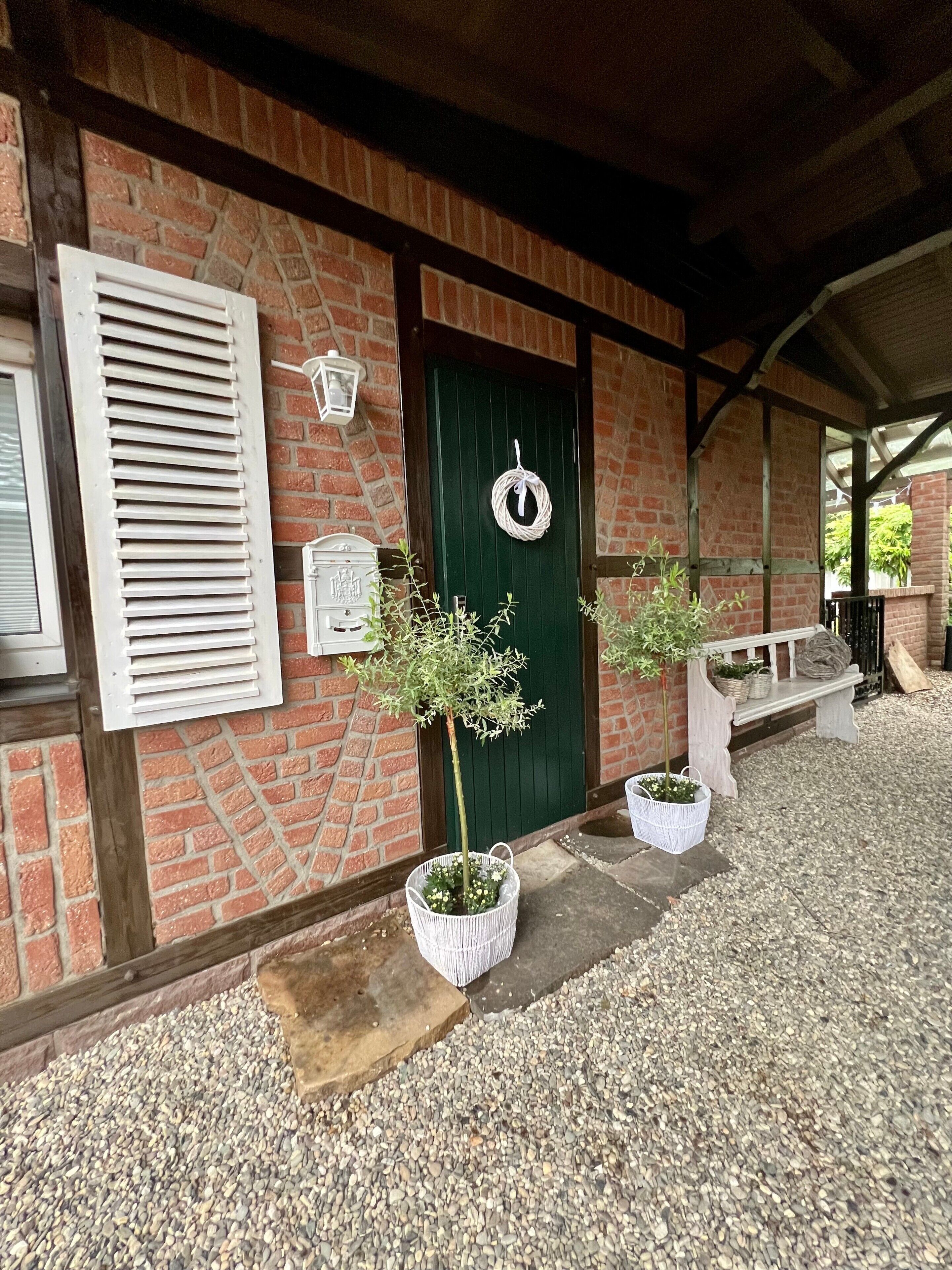 Un edificio de ladrillo con puerta verde, contraventanas blancas y un banco. Hay dos macetas con plantas en la entrada.