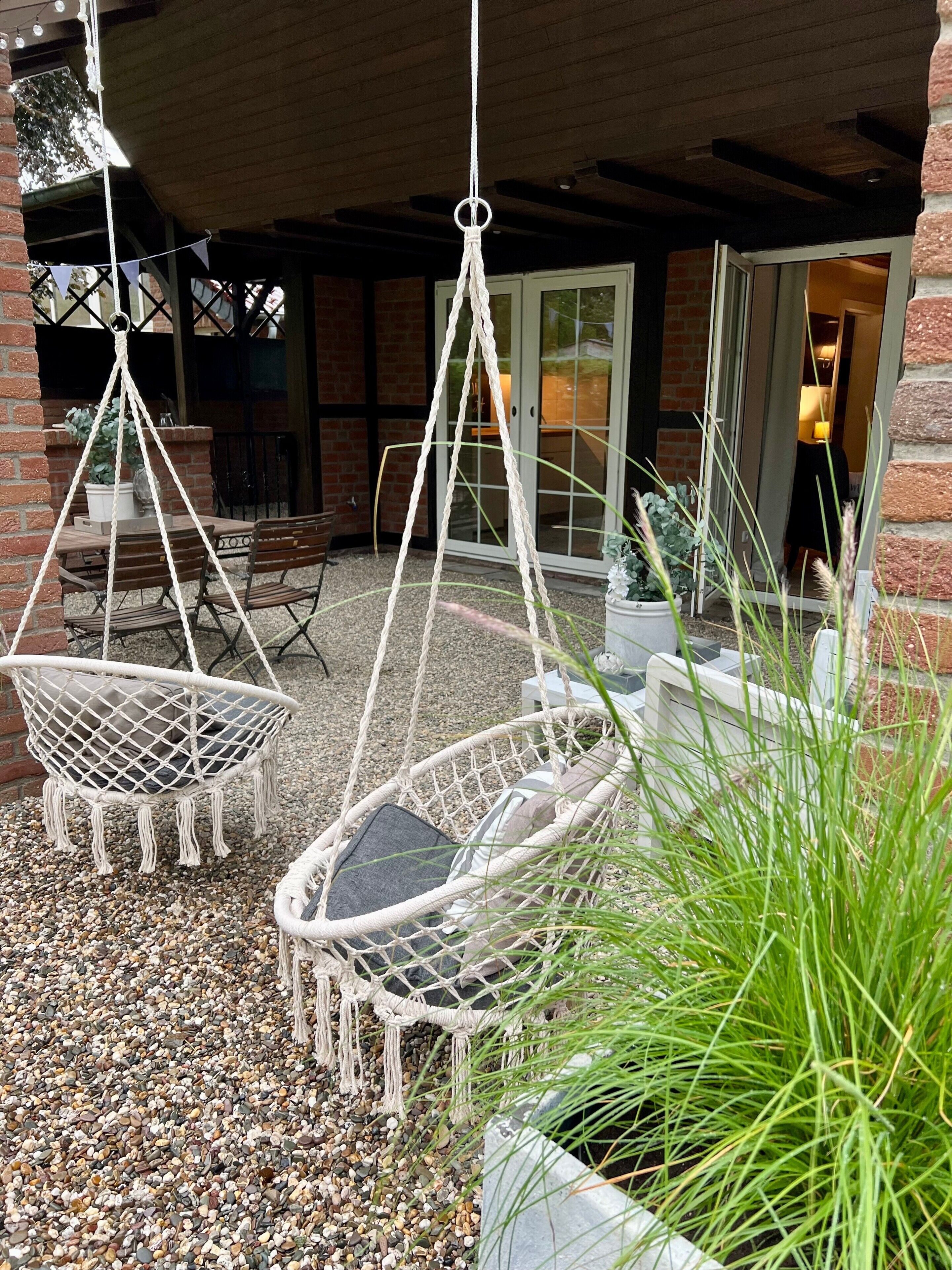 A covered outdoor seating area with two white wicker chairs and a table, surrounded by gravel and potted plants.