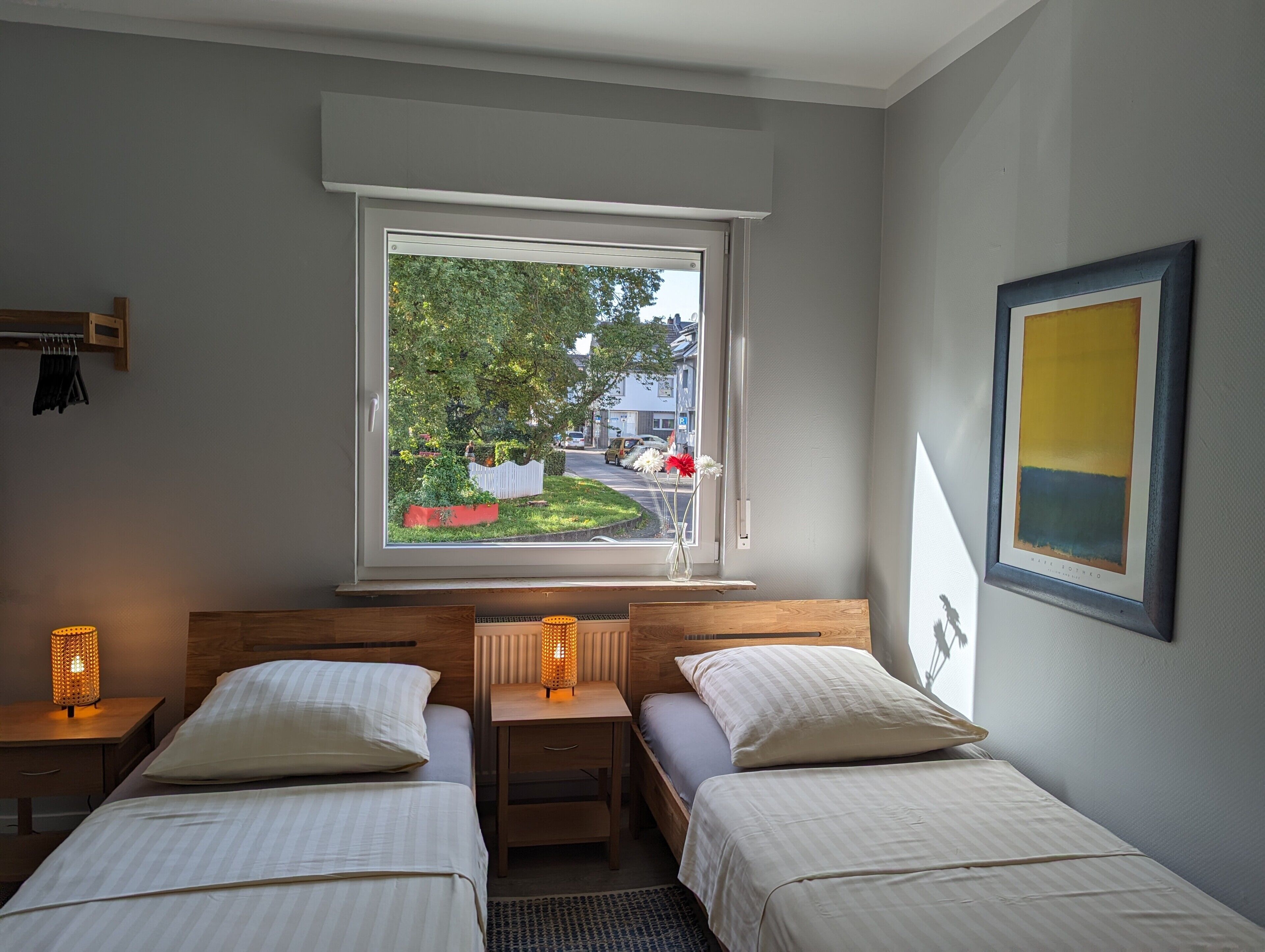 Een slaapkamer met twee bedden, een raam met uitzicht op een tuin en straat, een nachtkastje met een lamp, en een lijst met een afbeelding aan de muur.