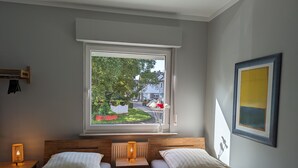 A bedroom with two beds, a window with a view of a garden and street, a bedside table with a lamp, and a framed picture on the wall. - Holiday Apartment "Goldgrube - Ground Floor" with Private Terrace, Shared Garden and Wi-Fi (Koblenz)