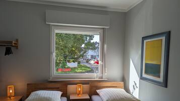 Een slaapkamer met twee bedden, een raam met uitzicht op een tuin en straat, een nachtkastje met een lamp, en een lijst met een afbeelding aan de muur.