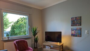 A living room with a television, a pink chair, a small table, and a window with a view of trees. - Holiday Apartment "Goldgrube - Ground Floor" with Private Terrace, Shared Garden and Wi-Fi (Koblenz)