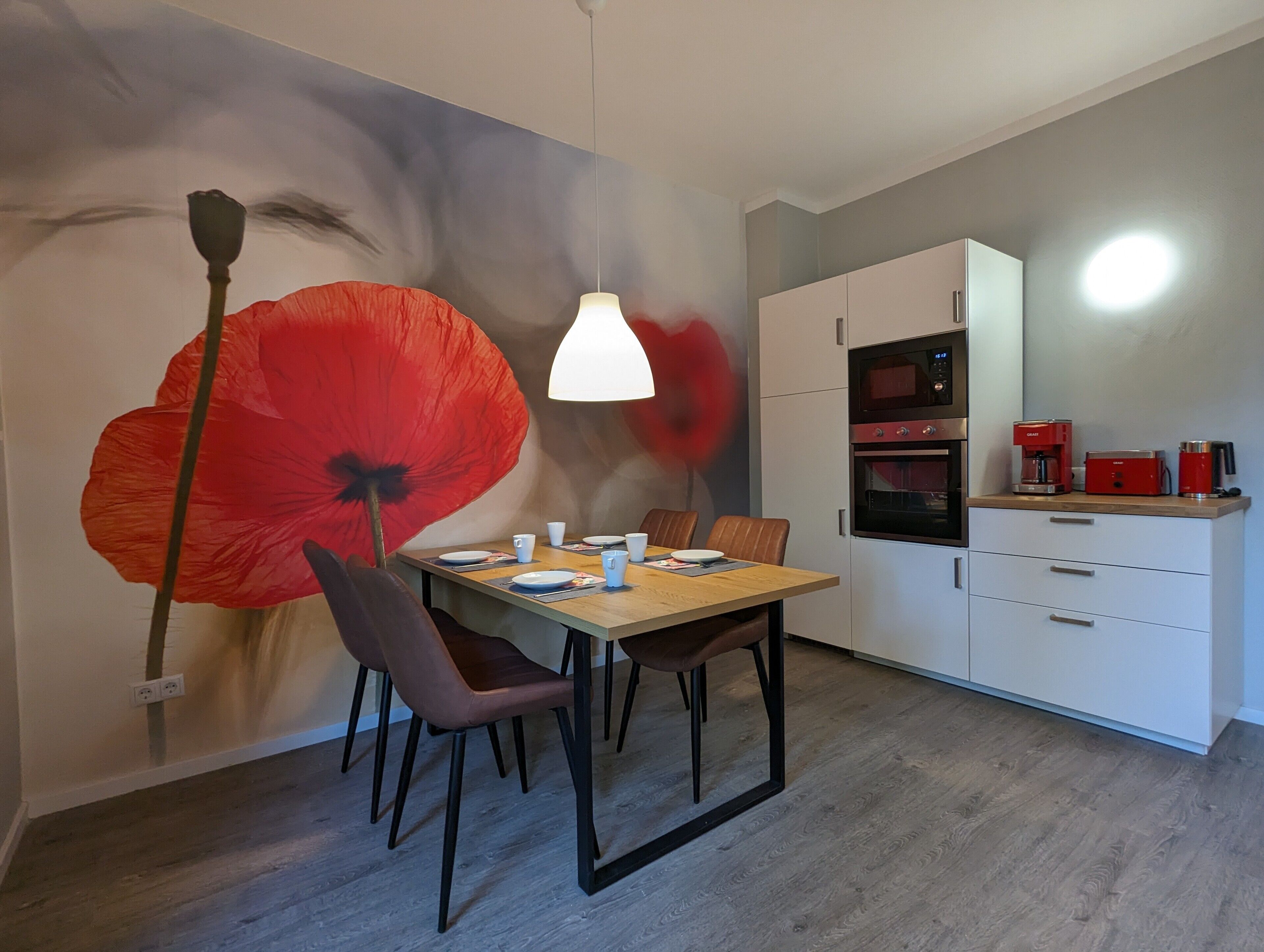 Een eetgedeelte met een tafel voor twee personen, een moderne keuken met apparatuur, en een grote muurschildering.