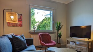 A modern living room with a sofa, armchair, coffee table, TV, and a window with a view of greenery.
