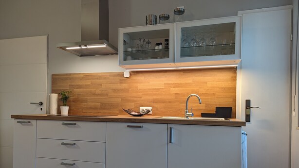 A modern kitchen with white cabinetry, a wooden backsplash, a stainless steel sink, and a glass-fronted upper cabinet.