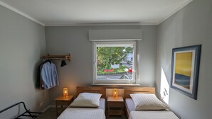 A bedroom with two beds, a window with a view of the street, a bedside table with a lamp, a bench, and a coat rack. - Holiday Apartment "Goldgrube Attic" with Wi-Fi (Koblenz)