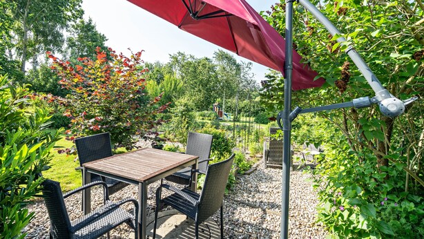 Un patio al aire libre con mesa y sillas, rodeado de vegetación y un paraguas rojo.