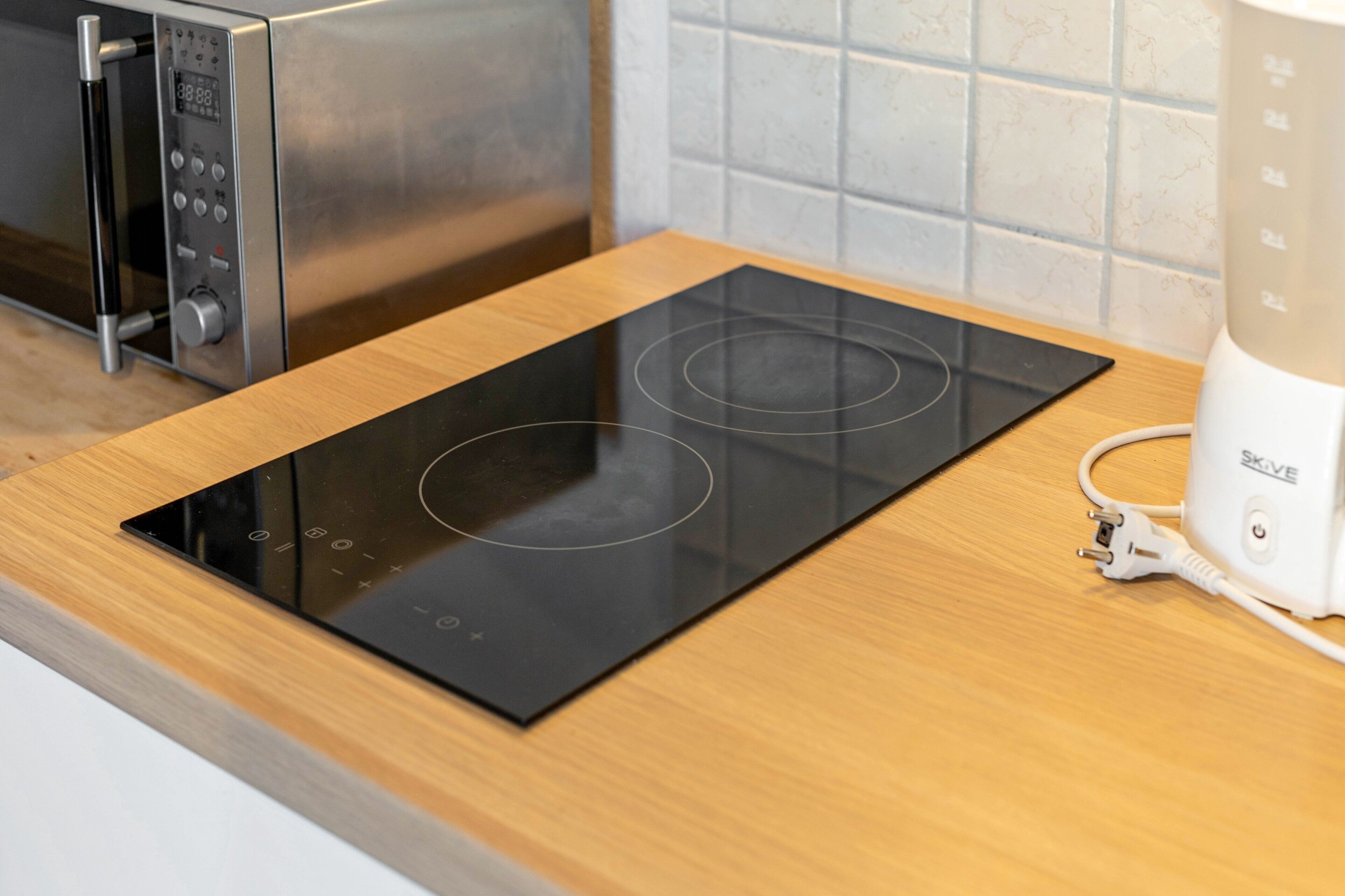 A kitchen with a black induction cooktop, a microwave, and a white blender on a wooden countertop.