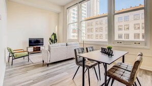 Una sala de estar moderna con sofá, mesa de comedor y sillas, televisor de pantalla plana y ventanas amplias con vista a la ciudad.