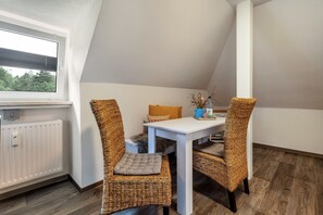 Une petite pièce triangulaire, avec une table blanche et deux chaises en osier, un radiateur et une fenêtre avec des stores.