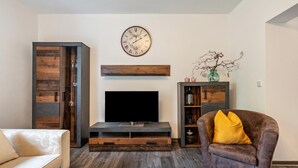 A living room with a flat-screen TV, wooden cabinets, a sofa, and a brown armchair with a yellow pillow. - Apartment 'Fewo Am Wald Erdgeschoss' with Shared Terrace, Shared Garden and Wi-Fi (Krauschwitz)
