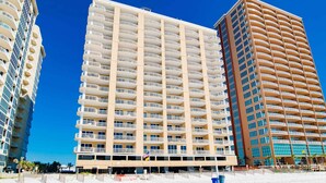 Un hotel moderno de gran altura con varios balcones, una playa al frente y un cielo azul despejado.