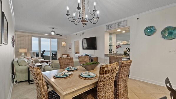 A dining area with wicker chairs, a wooden table, and a chandelier. There is a TV mounted on the wall and a sign that reads "ORANGE BEACH ALABAMA".