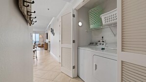 Lavandería con lavadora y secadora Whirlpool, repisa con cesteras y puerta con perchero.