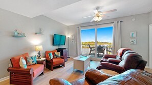 A living room with a sofa, armchair, and a small table. There is a ceiling fan, a TV, and a window with a view of the outdoors. - Unwind at Navy Cove 2106 – Relaxing Bay Views! (Gulf Shores)