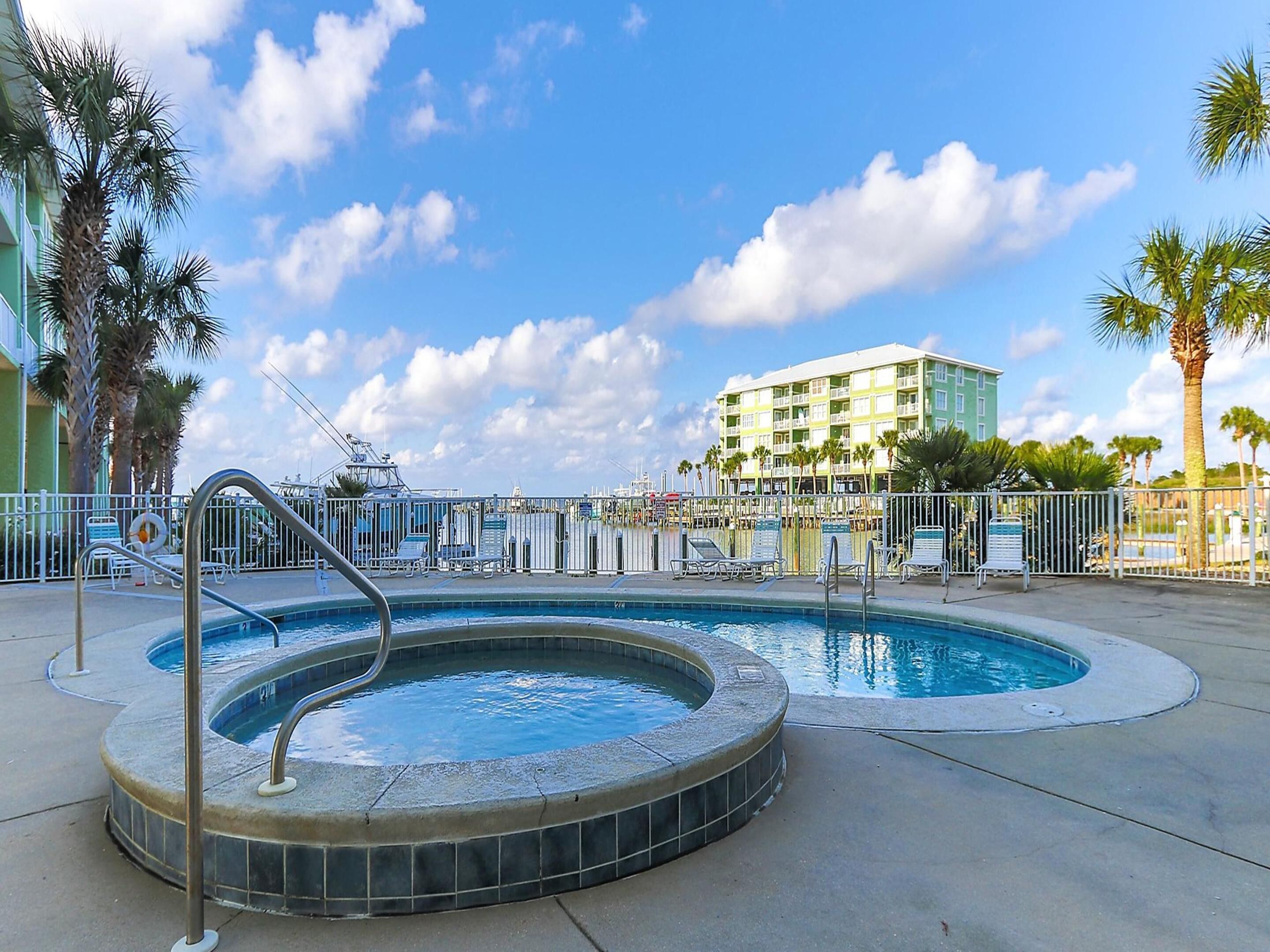 A swimming pool with a hot tub, surrounded by lounge chairs and palm trees, with a hotel building in the background.