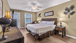 A bedroom with a bed, nightstands, a ceiling fan, a window with blinds, and a door. - Sea Oats F206 – Lagoon Views & Short Walk to Beach! (Gulf Shores)