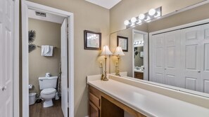 A bathroom with a toilet, sink, vanity, and a large mirror with lights. - Sea Oats F206 – Lagoon Views & Short Walk to Beach! (Gulf Shores)