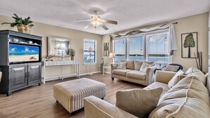 A living room with a sofa, ottoman, TV, console, and ceiling fan. - Sea Oats F206 – Lagoon Views & Short Walk to Beach! (Gulf Shores)