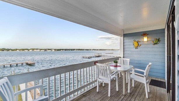 A covered outdoor deck with a view of the water, white chairs, a table, and decorative wall lights. - Sea Oats F206 – Lagoon Views & Short Walk to Beach! (Gulf Shores)