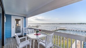 A covered outdoor area with a table and chairs, overlooking a body of water with docks and a distant shoreline. - Sea Oats F206 – Lagoon Views & Short Walk to Beach! (Gulf Shores)