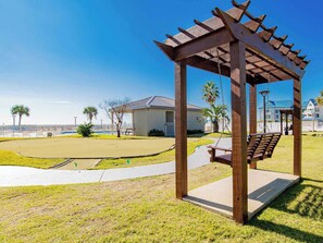 A wooden gazebo with a swing, a pool, and a building with a blue roof in the background. - Beachfront Living at Gulf Shores Plantation! (Gulf Shores)