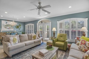 A living room with a ceiling fan, a sofa, a green armchair, and a coffee table with books.