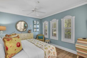 A bedroom with a bed, a chair, a dresser, a mirror, and three windows with shutters.