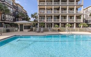 A pool area with a clear blue pool, surrounded by a fence, and adjacent to a multi-story building with balconies.