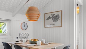 Un coin repas avec une table en bois, des chaises noires, une suspension orange, une carte de Skagen encadrée et une horloge murale.