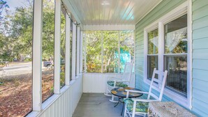 A covered porch with white chairs and a table, overlooking a street and trees. - 1 Mi to Beach: Sustainable Oak Island Home! (Oak Island)
