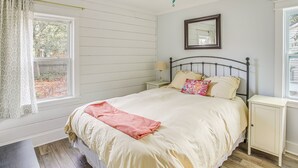 A bedroom with a bed, a nightstand, a ceiling fan, a window with curtains, and a small table. - 1 Mi to Beach: Sustainable Oak Island Home! (Oak Island)