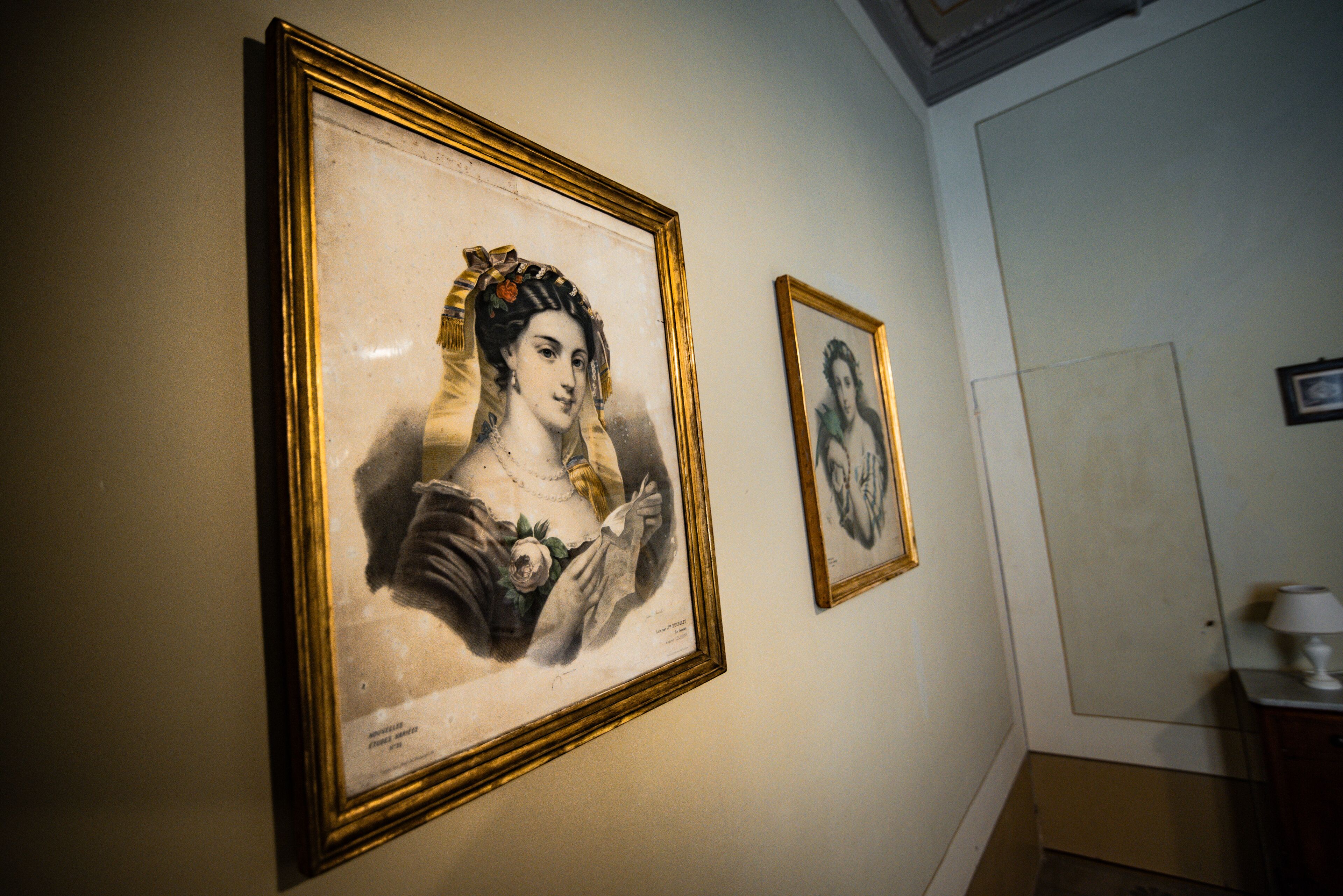 Two framed portraits on a wall in a room with a door and a small table with a lamp.