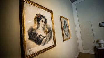 Two framed portraits on a wall in a room with a door and a small table with a lamp.