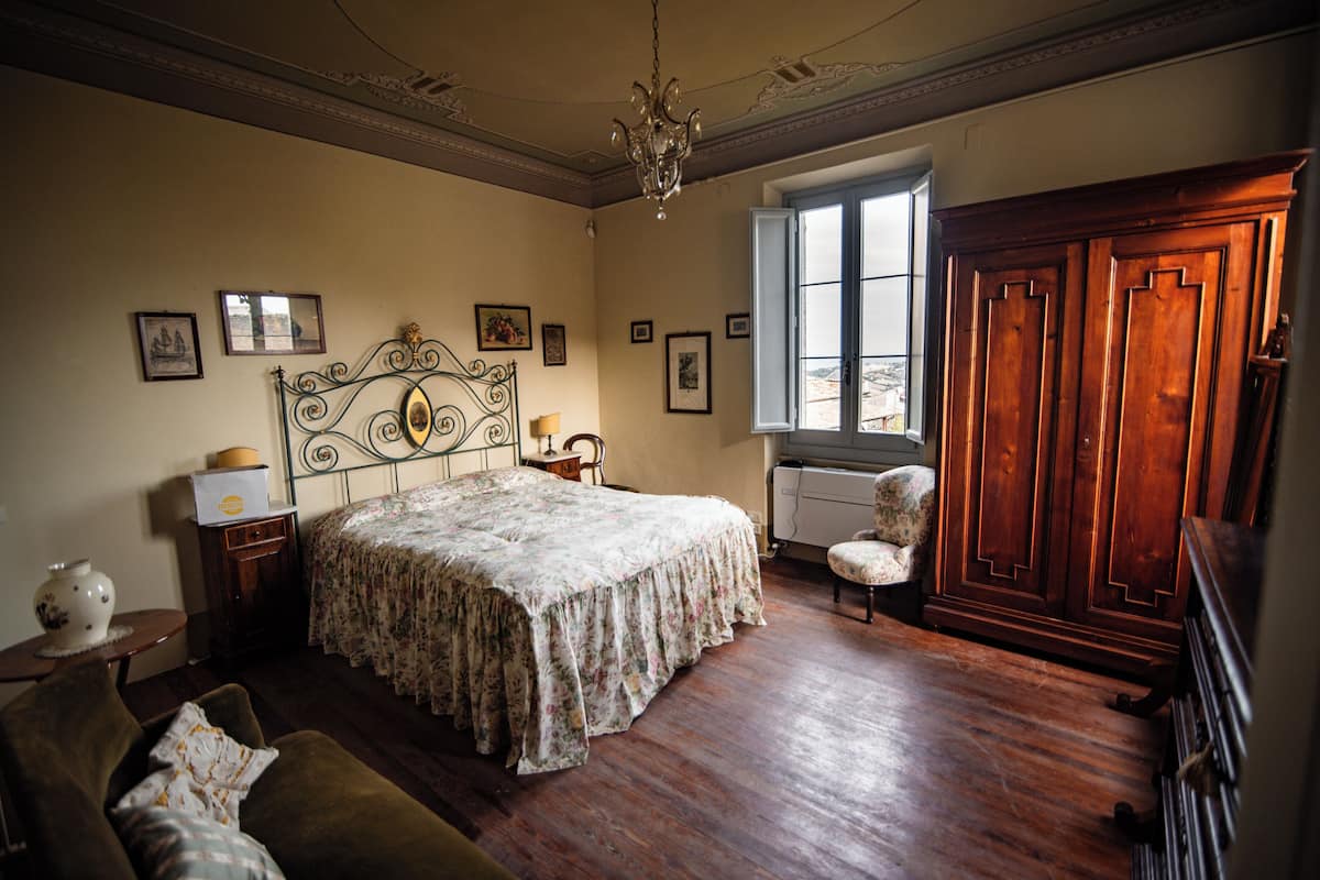 A bedroom with a large bed, a wooden wardrobe, a chandelier, and framed pictures on the walls.