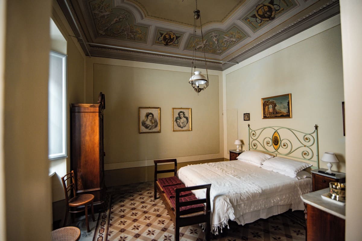 A bedroom with a large bed, two chairs, a wooden cabinet, and a ceiling with decorative artwork.