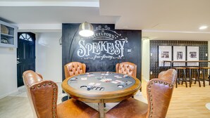 A room with a poker table, leather chairs, a black wall with 'SPEAK EASY' text, and a door.