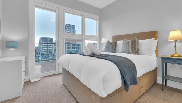 Une chambre moderne avec un grand lit, une table de chevet, une lampe et une vue sur la ville grâce à de grandes fenêtres.