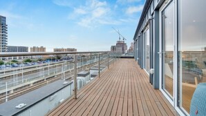 Une terrasse en bois avec des garde-corps en verre donnant sur un paysage urbain, avec notamment une voie ferrée et des immeubles.