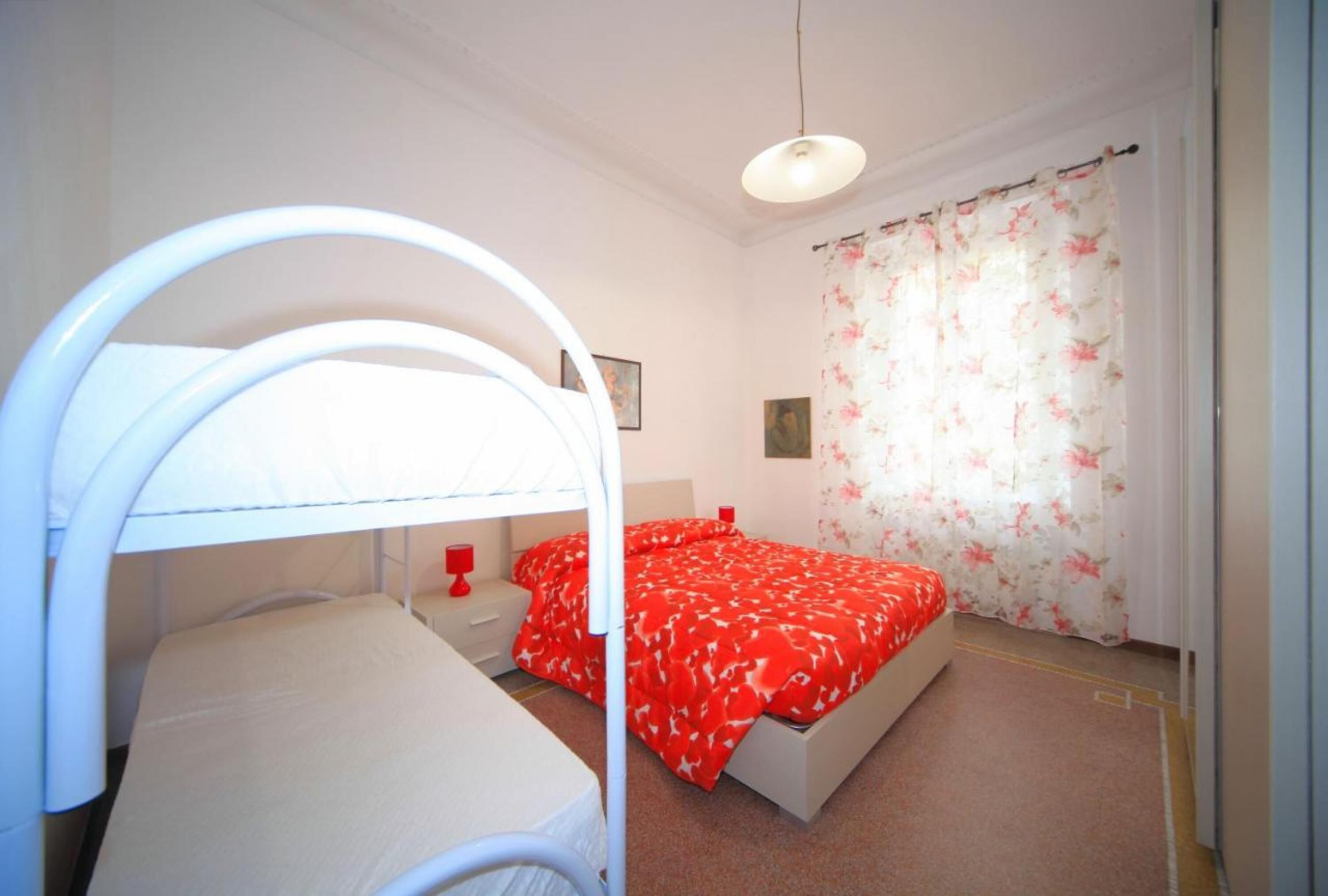 A bedroom with a bunk bed, a single bed, a bedside table with a red lamp, and a floral curtain.