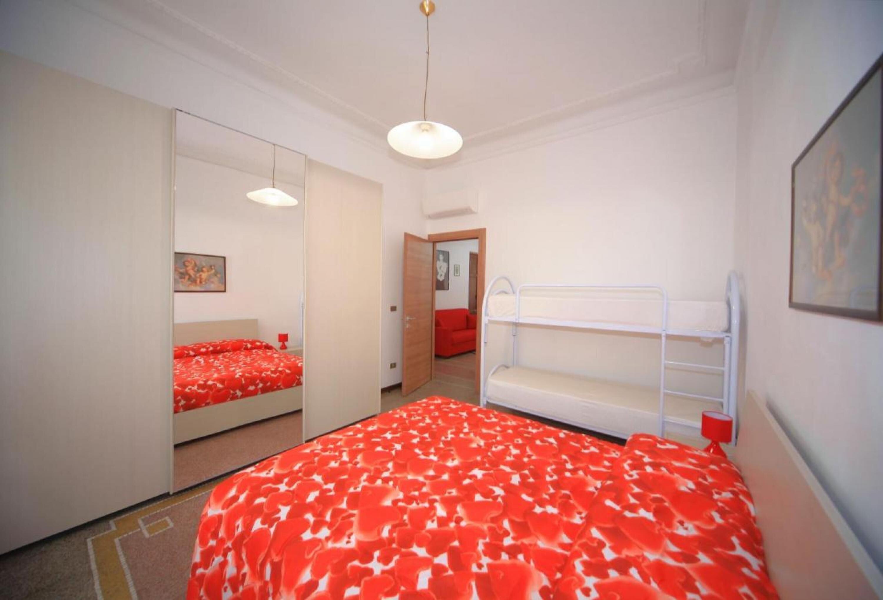 A bedroom with a bunk bed, a single bed with a red floral bedspread, a white wardrobe, a mirror, and a red lamp.