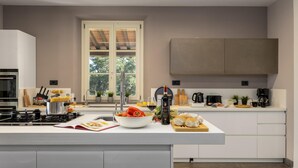 A modern kitchen with a central island, a built-in oven, and a window with a view of trees. - Villa Dada 10 Emma Villas (Rosignano Marittimo)