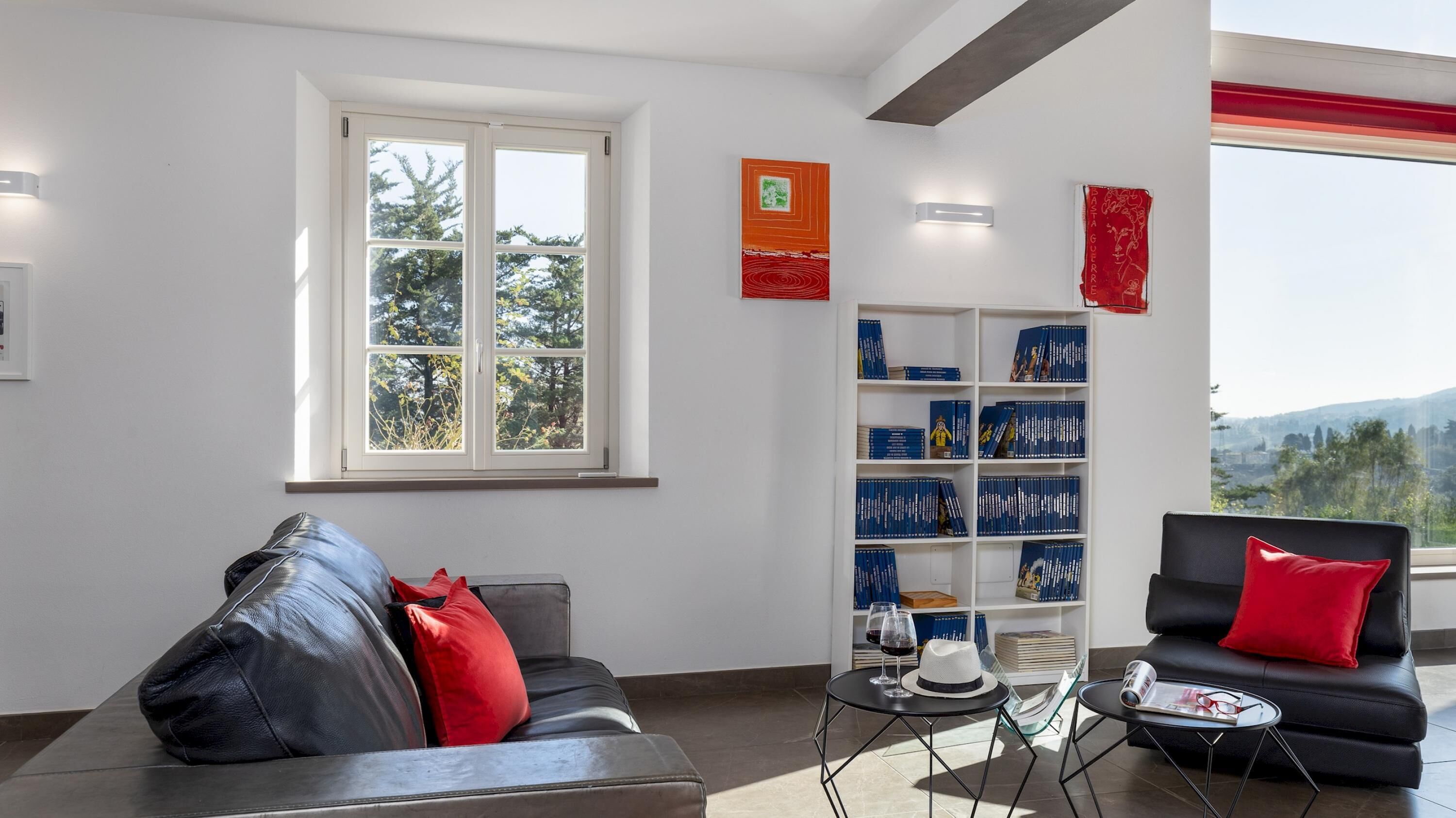 A modern living room with a leather sofa, a black armchair, a bookshelf, and a window with a view of trees and hills.
