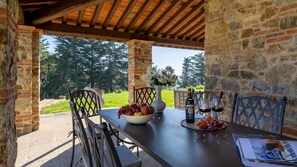 A covered outdoor dining area with stone walls, a table set with a bottle of wine, glasses, and a bowl of fruit.
